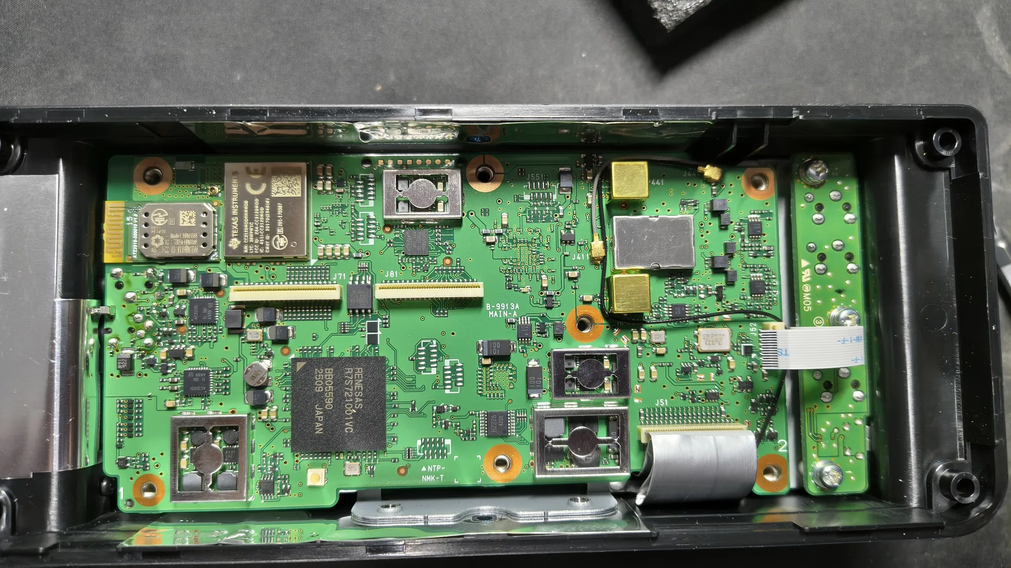 La IC-705 abierta, vista desde arriba: la placa principal verde dentro de la carcasa negra, con el módulo WiFi y Bluetooth de Texas Instruments arriba a la izquierda, el procesador Renesas en el centro, varios blindajes metálicos y el cable plano hacia la placa del frontal, a la derecha.